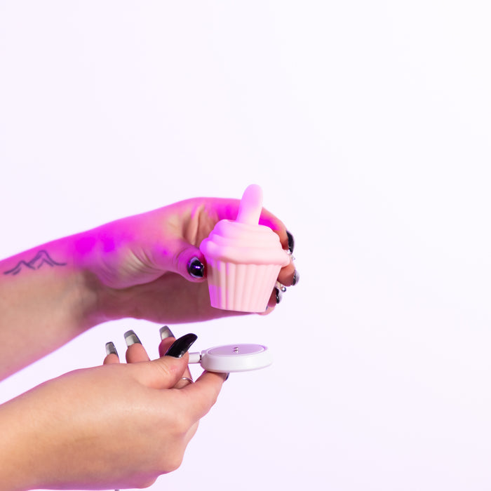 A person with long nails and a tattoo holds the pink Cake Eater Clitoral Stimulator Tongue Vibrator, shaped like a cupcake with a small top protrusion, in one hand and its white lid in the other against a white background.