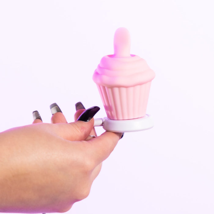 A hand with long, painted nails holds the Cake Eater Clitoral Stimulator Tongue Vibrator in pink, shaped like a cupcake with a soft swirl top, against a white background.