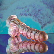 The Fantasy Ultra Textured Vibrating Remote Control Creature Dildo, a pink and pearlescent silicone toy with a flared base, rests on its side against a blue and purple abstract background, reflecting softly on a shiny surface.