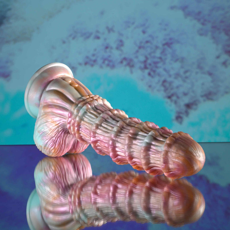 The Fantasy Ultra Textured Vibrating Remote Control Creature Dildo, a pink and pearlescent silicone toy with a flared base, rests on its side against a blue and purple abstract background, reflecting softly on a shiny surface.