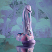 The Mythical Long Thick Vibrating Fantasy Dildo in Pastel Ombre is a smooth, abstract, pastel-colored toy with curved lines, displayed on a reflective surface against a marbled purple and blue background.