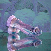 The Mythical Long Thick Vibrating Fantasy Dildo in Pastel Ombre, with its iridescent finish and phallic shape, rests on a shiny surface in front of a blurred abstract backdrop with pastel blue and purple tones.