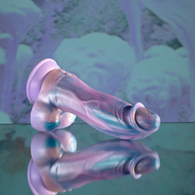 The Mythical Long Thick Vibrating Fantasy Dildo in Pastel Ombre, with its iridescent finish and phallic shape, rests on a shiny surface in front of a blurred abstract backdrop with pastel blue and purple tones.