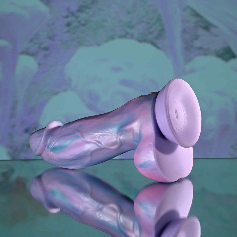 The Mythical Long Thick Vibrating Fantasy Dildo in Pastel Ombre, featuring a phallic shape and flared base, rests on a reflective surface with a blurred blue and purple abstract background.