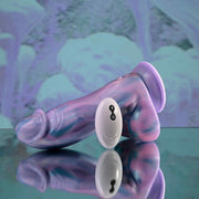 The Mythical Long Thick Vibrating Fantasy Dildo in Pastel Ombre rests on a reflective surface beside its small white remote, set against a soft purple and blue background.