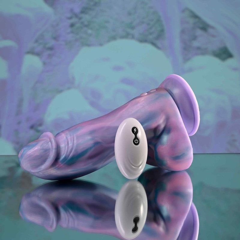 The Mythical Long Thick Vibrating Fantasy Dildo in Pastel Ombre rests on a reflective surface beside its small white remote, set against a soft purple and blue background.