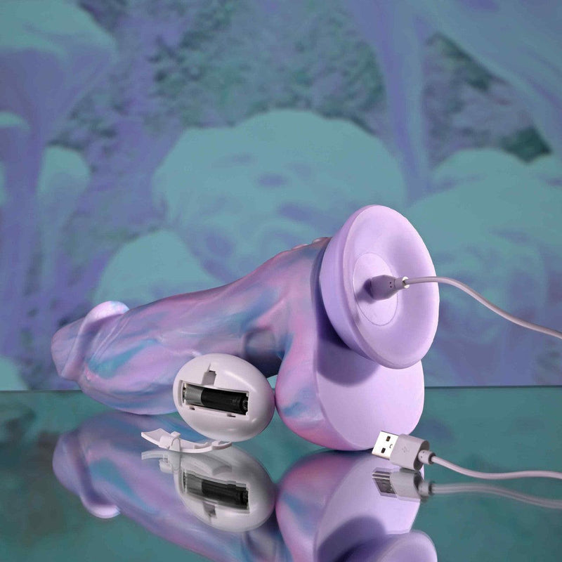 The Mythical Long Thick Vibrating Fantasy Dildo in Pastel Ombre is shown charging via USB cables on a reflective surface, set against a soft purple and blue abstract background.