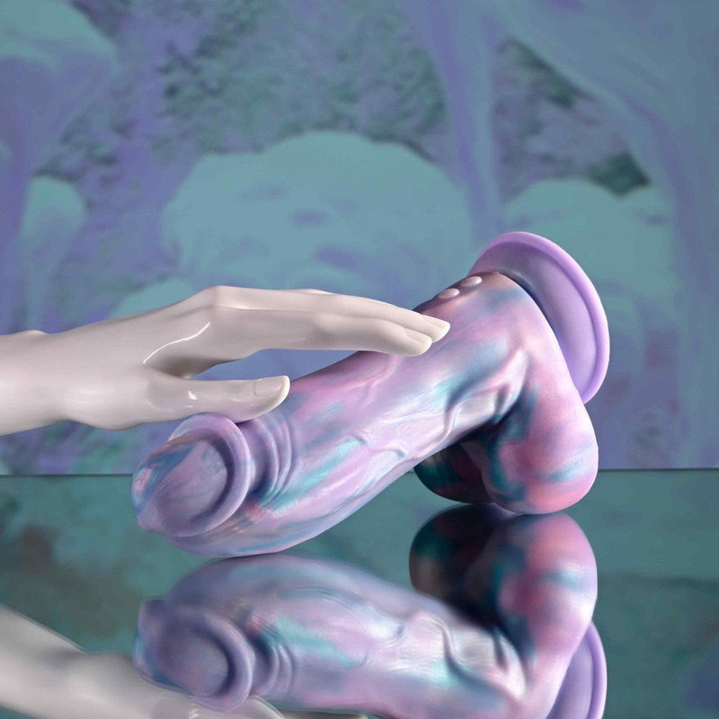 The Mythical Long Thick Vibrating Fantasy Dildo in Pastel Ombre rests on a reflective surface, with a mannequin hand posed above it. The background showcases soft, abstract designs in pastel hues that match the dildo's swirling colors.