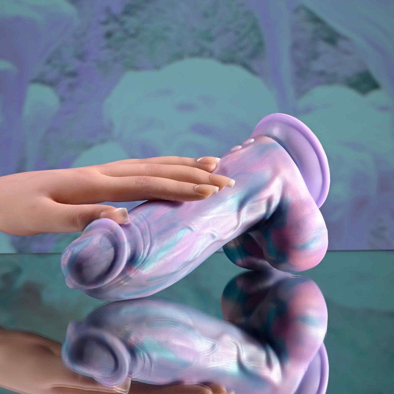 A manicured hand touches the Mythical Long Thick Vibrating Fantasy Dildo in Pastel Ombre, shaped like a phallus and placed on a reflective surface with a blurred, colorful background.