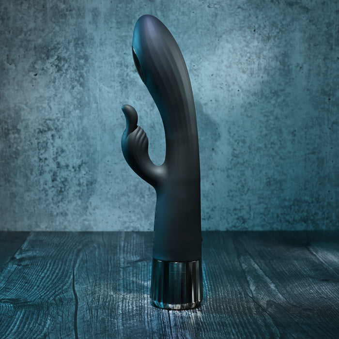 The Heat Up & Chill Dual Motor Rabbit with Heating & Cooling, a black silicone G-spot vibrator with a curved design and smaller extension, stands upright on a dark wooden surface against a textured gray background.