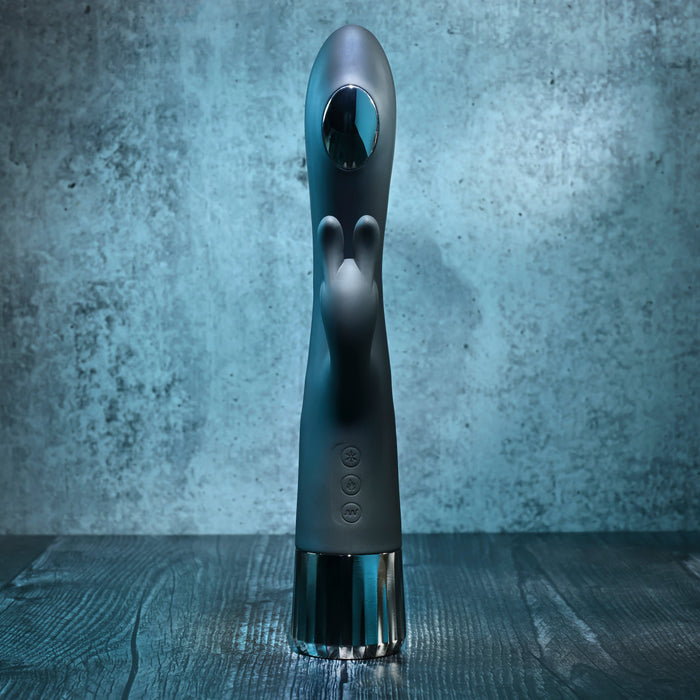 The Heat Up & Chill Dual Motor Rabbit with Heating & Cooling is a sleek black G-spot vibrator with two smaller protrusions, control buttons on the front, a metallic base, and is fully waterproof. It stands upright against a blue-gray background.