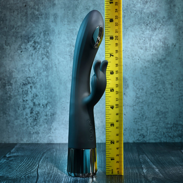 The Heat Up & Chill Dual Motor Rabbit with Heating & Cooling, a black silicone G-spot vibrator with a curved tip and external stimulator, stands upright on wood beside a yellow tape showing its 8-inch height against a textured gray wall.