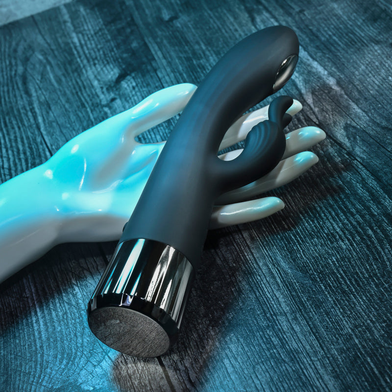 The Heat Up & Chill Dual Motor Rabbit with Heating & Cooling, in black and silver with a curved design and small extension, rests atop a glossy white mannequin hand on a dark wood surface.
