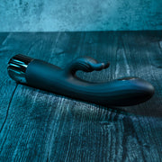 The Heat Up & Chill Dual Motor Rabbit with Heating & Cooling, a black silicone G-spot vibrator with curved shaft and external stimulator, rests on a textured dark wooden surface under blue lighting.