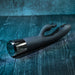The Heat Up & Chill Dual Motor Rabbit with Heating & Cooling, a black silicone vibrator with a metallic base, rests on a textured dark wooden surface with a blue-green tint.