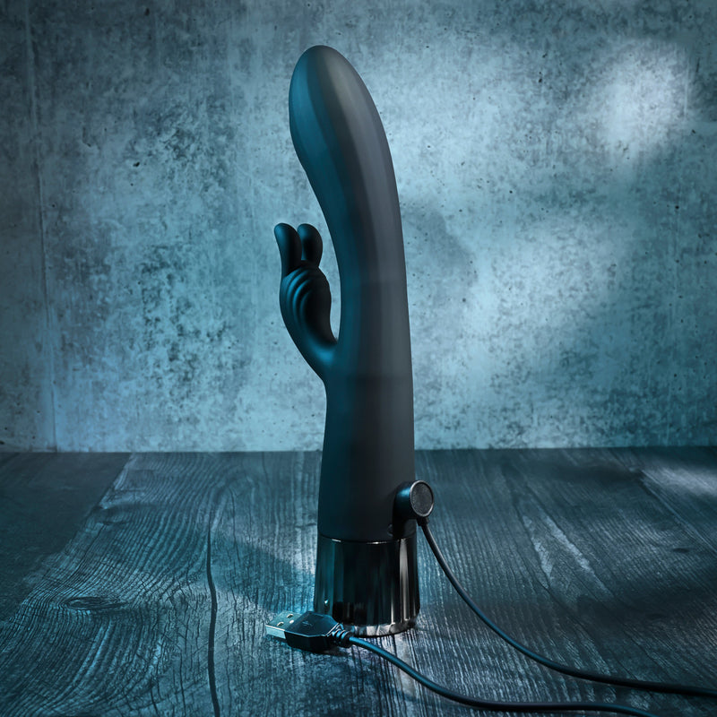 The Heat Up & Chill Dual Motor Rabbit with Heating & Cooling stands upright on a dark wooden surface, USB cable plugged in. The shadowy, blue-tinted background highlights this sleek black silicone vibrator and its waterproof design.