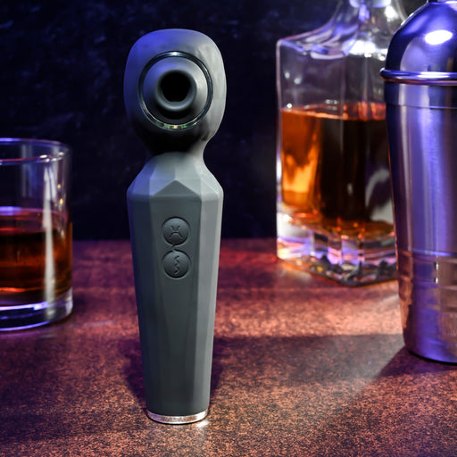 The Rainbow Sucker Wand + Clitoral Suction Vibrator stands upright on a bar counter beside a glass of whiskey, a decanter, and a cocktail shaker, all set against a dark and stylish backdrop.