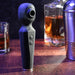 A black Rainbow Sucker Wand + Clitoral Suction Vibrator with buttons stands upright on a bar counter, surrounded by glasses and bottles of amber liquid, with a metal cocktail shaker in the background.