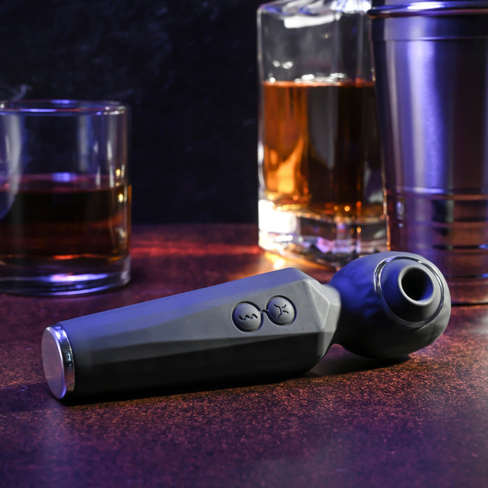 A Rainbow Sucker Wand + Clitoral Suction Vibrator with control buttons rests on a brown surface, while two glasses of whiskey, a decanter, and a metal cocktail shaker sit in the background.