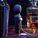 The Rainbow Sucker Wand + Clitoral Suction Vibrator, in black silicone, charges via USB on a bar counter amid a cocktail shaker, whiskey glass, and decanter against a dark background.