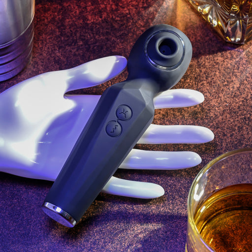 The Rainbow Sucker Wand + Clitoral Suction Vibrator, a sleek black dual-action vibe with two buttons, is displayed on a white mannequin hand on a brown textured surface beside a glass of amber liquid and a crystal container.