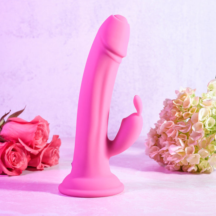 The Somebunny to Love Uncut Look Rabbit Vibrator, in pink with a suction base and clitoral stimulator, stands upright on a light surface among pink roses and hydrangeas against a pale backdrop.