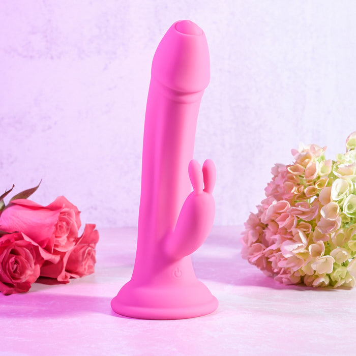 The Somebunny to Love Uncut Look Rabbit Vibrator, featuring a phallic shape and rabbit-shaped clitoral stimulator, stands upright among pink roses on the left and light hydrangeas on the right.