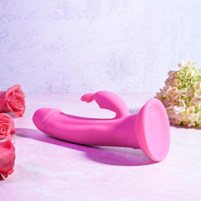 The Somebunny to Love Uncut Look Rabbit Vibrator, a pink curved waterproof silicone toy, rests on a light pink surface with pink roses and pale green hydrangeas nearby, set against a pastel textured background.