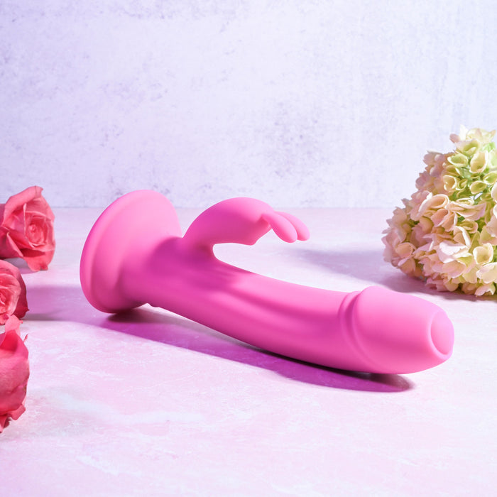 The Somebunny to Love Uncut Look Rabbit Vibrator, a pink dual motor toy with suction base and clitoral stimulator, is displayed on a pastel pink surface amid roses and light hydrangeas.