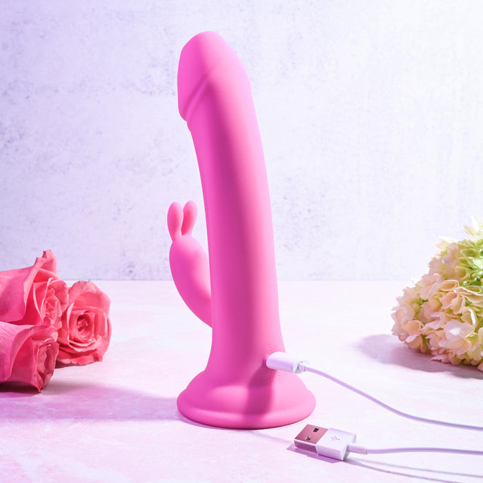 The Somebunny to Love Uncut Look Rabbit Vibrator in pink stands upright on a light surface with its USB charging cable attached, surrounded by pink roses and soft flowers against a pale, textured background.