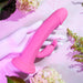 A white mannequin hand holds the Somebunny to Love Uncut Look Rabbit Vibrator, pink with a rabbit-shaped clitoral stimulator, surrounded by flowers and leaves on a light pink surface.