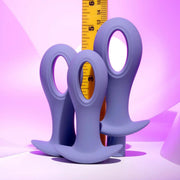 The Hole Punch Vibrating 3 Piece Anal Training Set, featuring purple silicone plugs with round handles and a sleek, hollow design, is shown in front of a yellow measuring tape against a pink-to-purple gradient background.