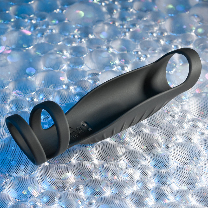 The Rocketeer Vibrating Penis Enhancer Sheath, a black body-safe silicone sheath with three loops, rests on a textured, iridescent bubble-patterned surface.