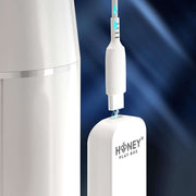 Close-up of the Blanche Thrusting Auto Blowjob Masturbator by Honey Play Box being connected to a USB-C cable, with blue arrows showing the direction, set against a dark, blurred background.