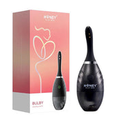 The Bulby Auto Spray Anal Douche System by Honey Play Box, a black automatic spray douche with USB rechargeable design, stands beside its pink box featuring white abstract outlines and branded product name.