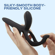 A hand holds the Pleasure Pivot Vibrating Cock Ring with Clit Stimulator, made of black body-friendly silicone, against a light blue background. Dark blue text above says, "Silky-Smooth Body-Friendly Silicone.