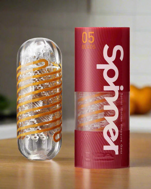 The Tenga Spinner Masturbation Cup 05 Beads, a clear cylindrical stroker with orange spiral ridges, is shown beside its red and transparent packaging labeled "Spinner 05 Beads" on a countertop with a blurred kitchen background.