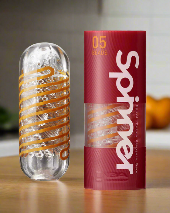 The Tenga Spinner Masturbation Cup 05 Beads, a clear cylindrical stroker with orange spiral ridges, is shown beside its red and transparent packaging labeled "Spinner 05 Beads" on a countertop with a blurred kitchen background.