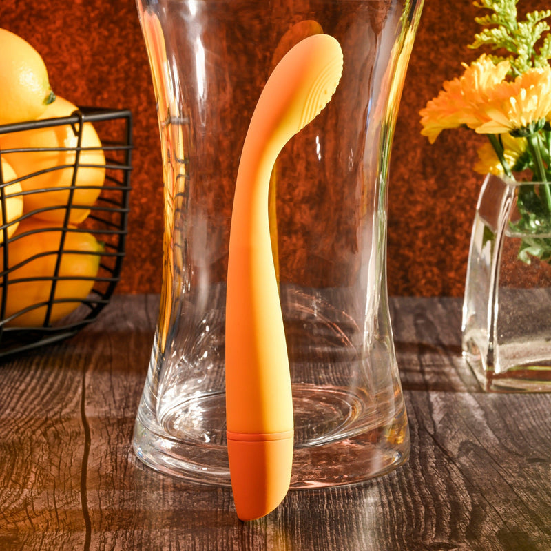 A yellow Lemon Squeeze Slim G-Spot Beginner Vibrator by Evolved Novelties with a curved head stands inside a clear vase on a wooden table. There is a black wire basket with lemons on the left and a clear square vase with yellow flowers on the right, all against a warm-toned background, resembling the allure of silky silicone designs.