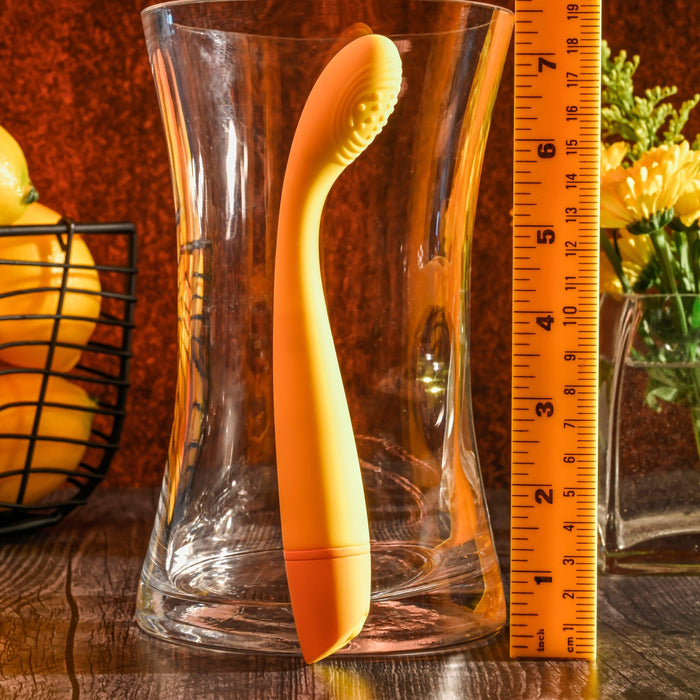 An orange Lemon Squeeze Slim G-Spot Beginner Vibrator from Evolved Novelties, crafted with silky silicone and featuring a curved handle, stands upright inside a glass vase. A yellow ruler is positioned next to the vase, measuring the height of the vibrator. In the background, there is a basket of lemons and a vase of yellow flowers.