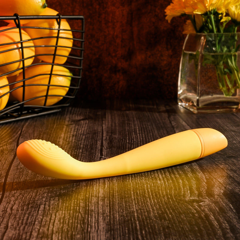 A yellow Lemon Squeeze Slim G-Spot Beginner Vibrator from Evolved Novelties, crafted from silky silicone and designed with a sleek curve, rests on a dark wooden surface. In the background stands a metal basket brimming with lemons and a glass vase showcasing vibrant yellow flowers. The setting exudes warmth and charm, creating an inviting rustic atmosphere.