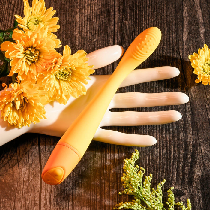 The Lemon Squeeze Slim G-Spot Beginner Vibrator in yellow by Evolved Novelties is elegantly placed on a white mannequin hand, surrounded by yellow flowers. The scene, set on a dark wooden surface, creates a striking contrast with the brightly colored flowers and the waterproof massager's textured silicone surface designed for G-spot stimulation.