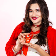 A smiling woman with long brown hair, red lipstick, and bold eye makeup holds the Little Red Discreet Powerful Bullet Vibrator. She wears a white dress, red shawl, red choker, and silver rings against a white background.