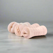 The M for Men 3-Pack Self-Lubricating Vibrating Masturbation Sleeve Kit - Vanilla displays three realistic, detailed silicone sleeves on a gray surface, each designed to provide a lifelike experience for adult pleasure.