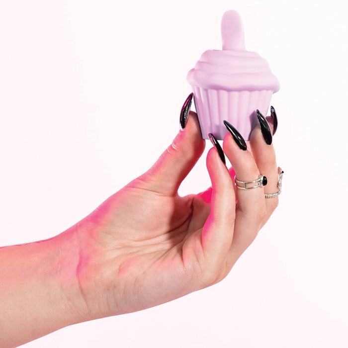 A hand with long black nails and silver rings holds the Cake Eater Clitoral Stimulator Tongue Vibrator in pink, shaped like a cupcake with a swirl top, against a white background.