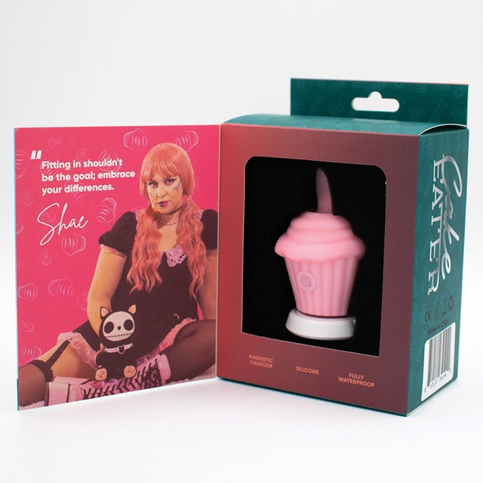 The Cake Eater Clitoral Stimulator Tongue Vibrator - Pink sits boxed beside an open flap showing a person with long pink hair in a black dress holding a plush toy. The flap reads, "Fitting in shouldn't be the goal; embrace your differences.