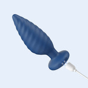 The Noah App-Enabled Rotating Butt Plug, a blue ribbed cone-shaped anal toy with a flared base, is shown being charged with a white USB cable connected to its bottom.