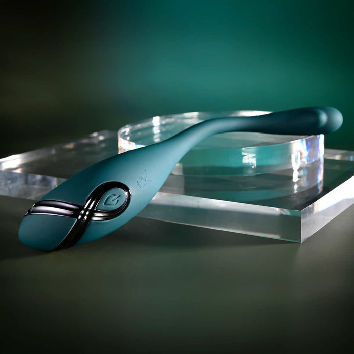 The Pinpoint Perfection Flexible Slim Vibrator, featuring a sleek teal design with a black accent, is displayed on a clear acrylic platform against a dark green gradient background.