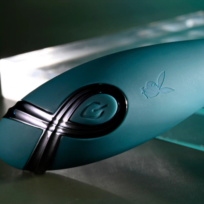 A close-up of the Pinpoint Perfection Flexible Slim Vibrator in sleek teal, featuring a black power button and Playboy bunny logo, set against a glass and metal background with dramatic lighting.