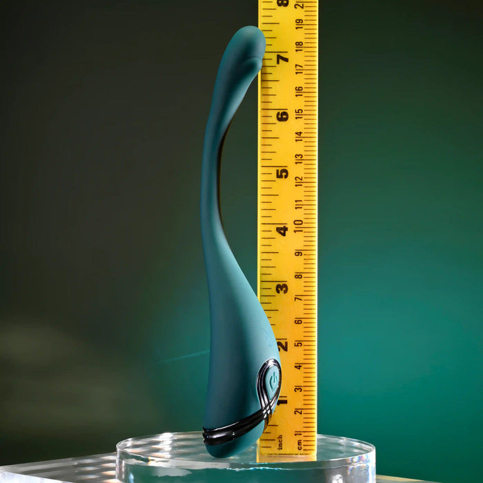 The Pinpoint Perfection Flexible Slim Vibrator, a curved teal toy, stands upright in a clear dish next to a yellow measuring tape showing its height, all set against a green background.